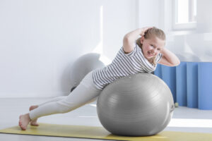 Smiling girl in striped shirt doing exercise on grey ball during rehabilitation in hospital room